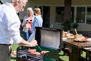 Smiling senior man making barbecue at outdoor grill while women drinking wine behind